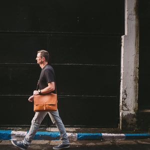 A man walking down a street with a large caramel brown leather messenger style bag over one shoulder. 